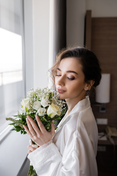 young woman with engagement ring on finger standing in white silk robe and looking at floral bridal bouquet next to window in hotel suite, special occasion, bride on wedding day - Photo, Image