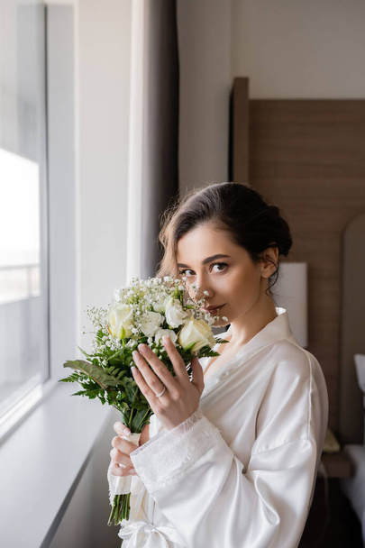 young bride with engagement ring on finger standing in white silk robe and smelling bridal bouquet next to window in hotel suite, special occasion, wedding day - Photo, Image