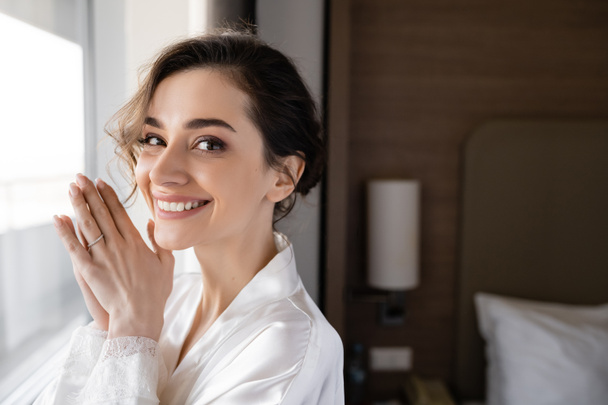 портрет очаровательной молодой женщины с обручальным кольцом на пальце, стоящей в белом шелковом халате и смотрящей в камеру в современном номере отеля, особый случай, невеста в день свадьбы - Фото, изображение