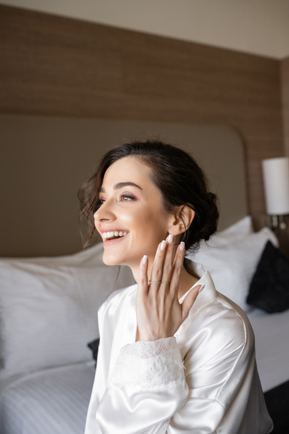 excited young woman with brunette hair in white silk robe showing engagement ring on finger and smiling in hotel room on wedding day, special occasion, young bride - Photo, Image