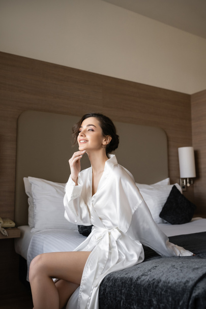 happy young bride with brunette hair sitting in white silk robe on comfortable bed and looking away in hotel suite on wedding day, special occasion, young bride - Fotografie, Obrázek