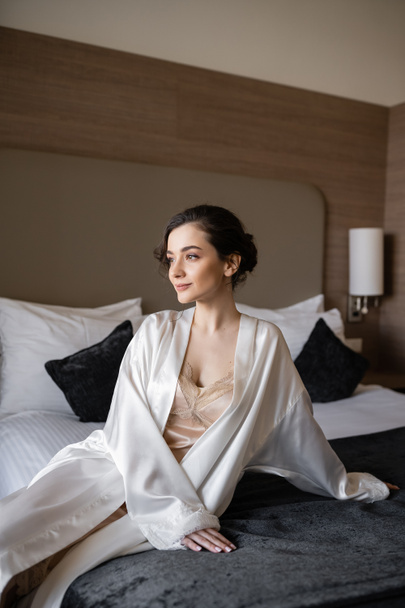 urocza panna młoda z brunetką włosy i makijaż ślubny siedzi w białym jedwabnym szlafroku na wygodnym łóżku i patrząc w apartamencie hotelowym przed jej ślubem, specjalna okazja  - Zdjęcie, obraz