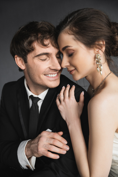 portrét nevěsty s brunetovými vlasy, elegantními šperky a bílými šaty objímající rameno šťastného ženicha v klasickém černém obleku s kravatou v moderním hotelovém apartmá po svatebním obřadu  - Fotografie, Obrázek