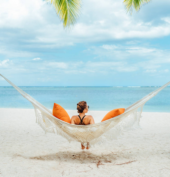 Young woman relaxing in wicker hammock on the sandy beach on Mauritius coast and enjoying wide ocean view waves. Exotic countries vacation and mental health concept image. - Fotoğraf, Görsel