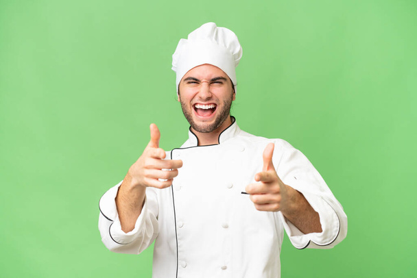 Молодой красивый шеф-повар на изолированном фоне, указывая на фронт и улыбаясь - Фото, изображение