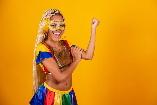 beautiful black Brazilian woman, dressed in carnival clothes, mask. yellow background. holding biceps, showing strength. - Valokuva, kuva