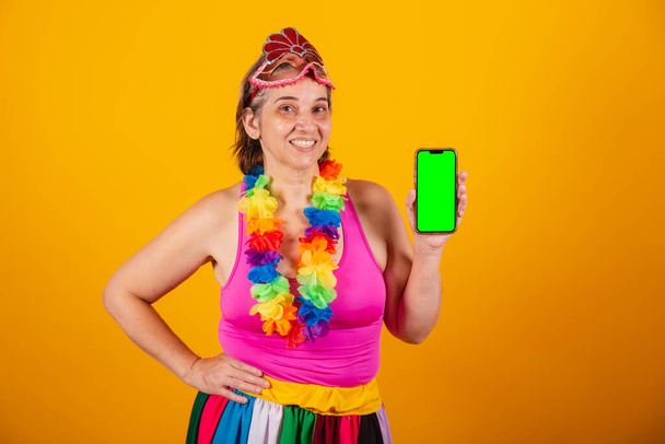 Adult woman in carnival clothes, smiling at camera, holding smartphone with green screen. - Foto, Imagem
