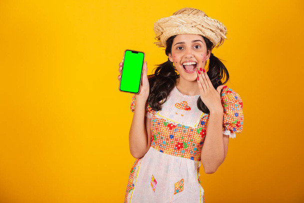 Piękna Brazylijka, z ubraniami festa junina, trzymając smartfon z zielonym ekranem. - Zdjęcie, obraz