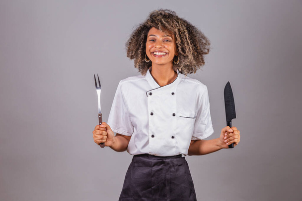 Young black Brazilian woman, cook, masterchef, wearing restaurant uniform. holding fork and knife for barbecue. Meat cutting. - Foto, afbeelding