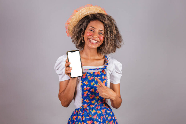 Young black Brazilian woman, with country clothes, dressed for Festa Junina. holding smartphone with white screen for ads, mobile apps. - Photo, Image