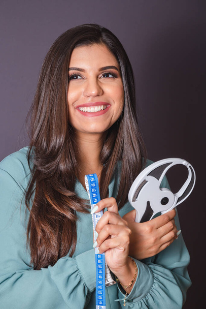 Horizontal photo. Beautiful Brazilian woman, with casual clothes, Jeans and green shirt. nutritionist holding measuring tape and adipometer - Foto, afbeelding
