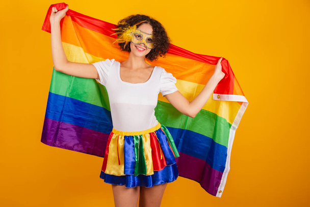 Beautiful Brazilian woman, dressed in carnival clothes, colorful skirt and white shirt. wearing mascara, holding lgbt flag. - Foto, Bild