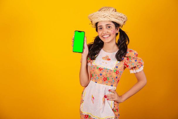 Beautiful Brazilian woman, with festa junina clothes, holding smartphone with green screen. - Foto, afbeelding