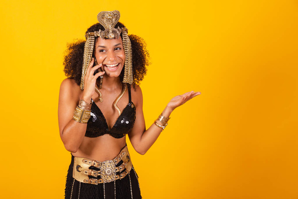 Beautiful black Brazilian woman, in Cleopatra carnival clothes, talking with smartphone, voice call. - Photo, image