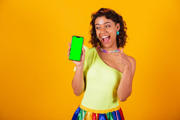 beautiful afro american brazilian woman, in carnival clothes, holding smartphone with green screen in chroma. - Photo, image