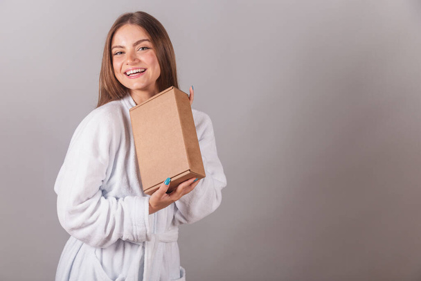 Brazilian blonde woman showing wonderful hair. dressed in a robe. holding medium cardboard box with cosmetics. - Photo, Image