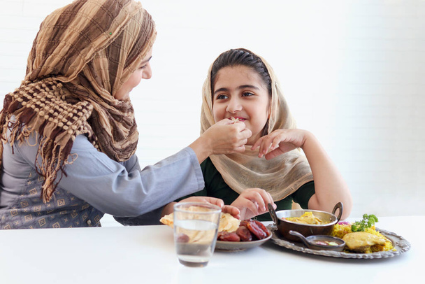 Adorable Muslim happy girl sits at kitchen table, kid with hijab enjoy eating traditional Islamic halal food with mother hand on white background, mom feeding child daughter, warm love in family. - Foto, Imagem