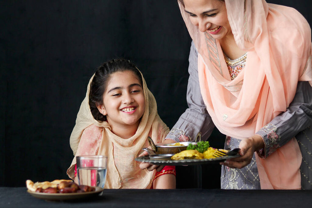 Очаровательная улыбающаяся пакистанская мусульманка с красивыми глазами, сидящая за кухонным столом, мать с хиджабом, подающая исламскую халяльную еду на тёмном чёрном фоне, мама заботится о дочери. семейная теплота любви - Фото, изображение