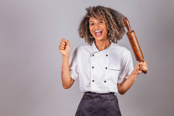 Young black Brazilian woman, cook, masterchef, wearing restaurant uniform. holding wooden rolling pin for preparing pasta, bread and pizza. - Foto, Bild