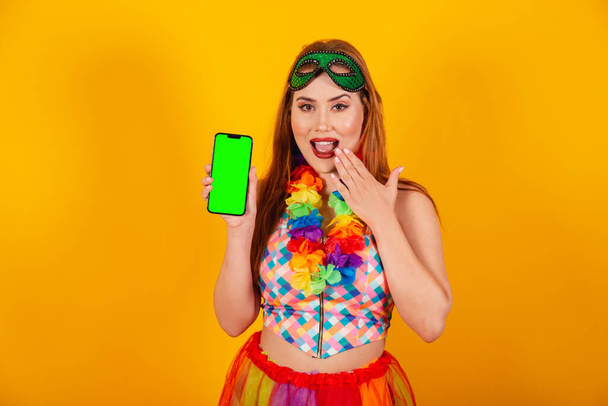 Brazilian redhead, in carnival clothes, with a necklace of flowers around her neck, showing a smartphone with a green chroma screen. - Foto, immagini