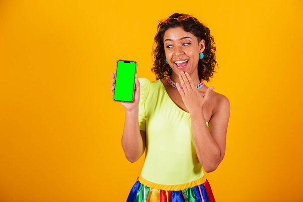 όμορφη αφροαμερικανίδα βραζιλιάνα, με ρούχα καρναβαλιού, κρατώντας smartphone με πράσινη οθόνη σε chroma. - Φωτογραφία, εικόνα