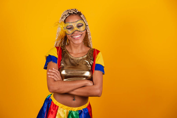 beautiful black Brazilian woman, dressed in carnival clothes, mask. yellow background. crossed arms. - Фото, зображення
