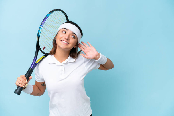 Genç tenisçi kadın mavi arka planda izole edilmiş bir şekilde elini kulağına koyarak bir şeyler dinliyor. - Fotoğraf, Görsel