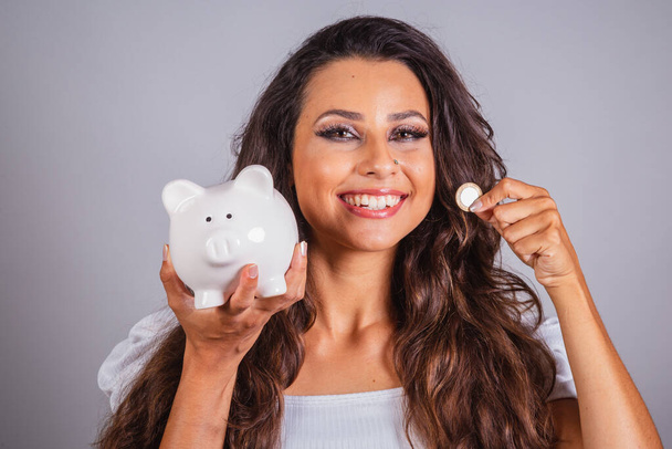 Brazilian woman, brown hair, holding piggy bank and coin. Finance. pension. - Zdjęcie, obraz