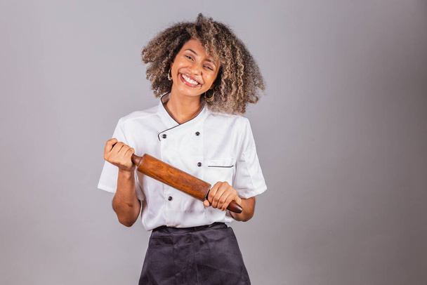 Young black Brazilian woman, cook, masterchef, wearing restaurant uniform. holding wooden rolling pin for preparing pasta, bread and pizza. - 写真・画像