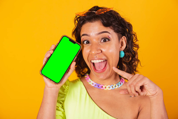 beautiful afro american brazilian woman, in carnival clothes, holding smartphone with green screen in chroma. - Photo, Image