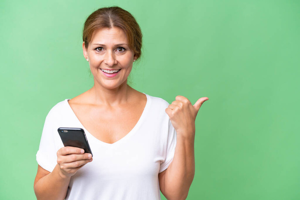 Middle-aged caucasian woman over isolated background using mobile phone and pointing to the lateral - Φωτογραφία, εικόνα