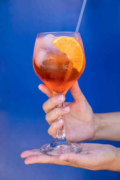 Um copo de Aperol spritz cocktail em mãos femininas sobre um fundo azul. Vertical. Close-up. - Foto, Imagem