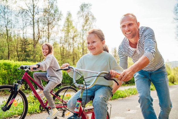 Lächelnder Vater mit zwei Töchtern beim Spaziergang im Freien. Er brachte jüngeren Mädchen das Fahrradfahren bei. Sie genießen die Zweisamkeit im sommerlichen Stadtpark. Bild einer glücklichen Kindheit. - Foto, Bild