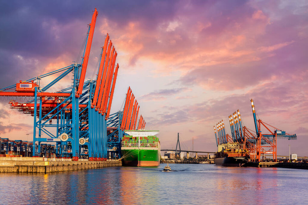 Γραφικό μπροστινό γιγαντιαίο φορτηγό πλοίο μεταφοράς φορτίου φόρτωση λιμάνι λιμάνι Αμβούργο γερανοί λιμάνι ζεστό δραματικό ουρανό βράδυ φως ηλιοβασίλεμα. Παγκόσμιο εμπορικό εμπόριο ναύλωσης ναύλωσης ναυτιλιακή εφοδιαστική υπόβαθρο. - Φωτογραφία, εικόνα