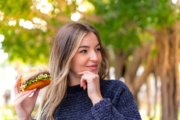 Genç ve güzel Romen kadın elinde hamburgerle dışarıda bir fikir üretiyor ve bir tarafa bakıyor. - Fotoğraf, Görsel