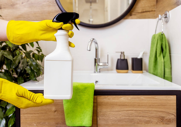 close-up of a female hand in a protective glove holding white bottle, cleaning agent mockup, space for logo and label. Bathroom background. The concept of hygiene, cleanliness and cleaning in house - Photo, image