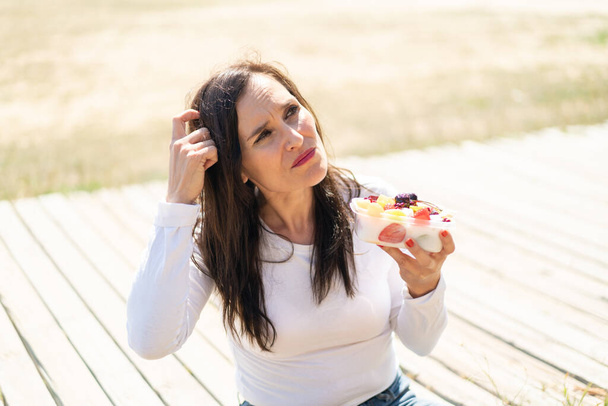 Жінка середнього віку тримає миску фруктів на відкритому повітрі, сумнівається і з плутаниною вираження обличчя
 - Фото, зображення