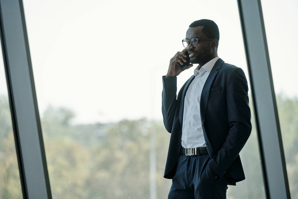 Young African American chief executive officer in formalwear speaking on mobile phone while standing by large office window - Foto, afbeelding