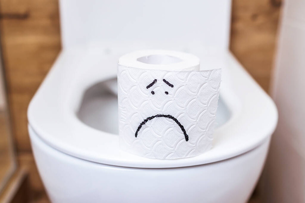 zbliżenie rolki białego papieru toaletowego z pomalowaną smutną twarzą stoi na toalecie. Koncepcja złego nastroju, problemy zdrowotne, zaparcia, hemoroidy, problemy jelitowe. Higiena osobista, samodzielna opieka - Zdjęcie, obraz