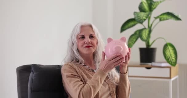 Elderly woman shakes piggy bank and listens to sound inside. Budget spending and the creation of savings and effective investment - Footage, Video