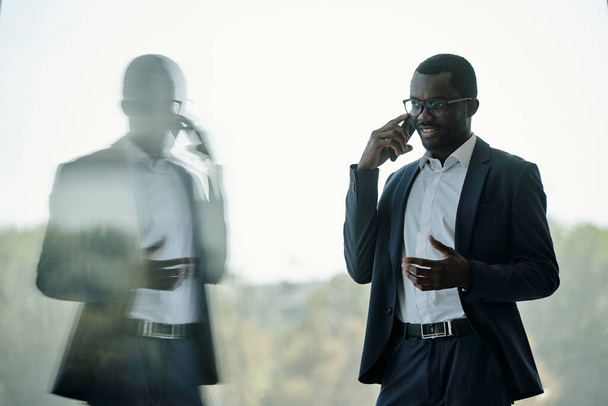 オフィスの窓の前に立って耳で携帯電話で若い成功したアフリカ系アメリカ人のビジネスマンや起業家 - 写真・画像