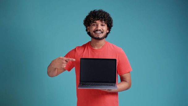 Портрет позитивної радісної брюнетки латинської або арабської людини з бородою в білій футболці, що тримає ноутбук з порожнім екраном і посміхається на камеру, інтернет-рекламі. студійний знімок ізольований на синьому студійному фоні
. - Фото, зображення