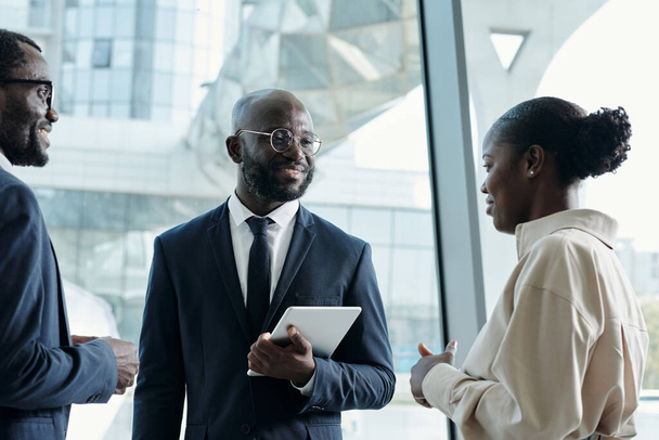 Ευτυχισμένος νεαρός μαύρος επιχειρηματίας με επίσημο ένδυμα κοιτάζοντας όμορφη γυναίκα συνάδελφο με λευκή μπλούζα κατά τη διάρκεια συζήτησης των ιδεών τους - Φωτογραφία, εικόνα