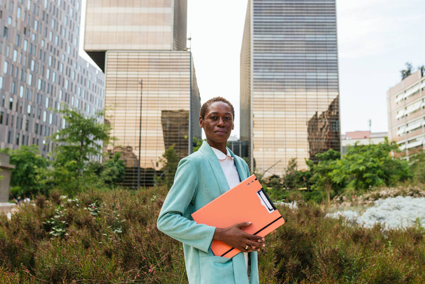 An African woman dressed in business attire holds a folder in her hand, looking confidently and serenely at the camera. She exudes empowerment and leadership with her short afro hair. The background - Фото, зображення