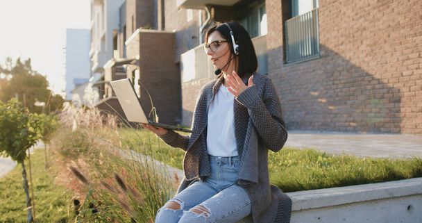 Beautiful young woman sitting in headphones with laptop computer in hands during studying remote lessons or working freelance speaking in camera at courtyard - Photo, image