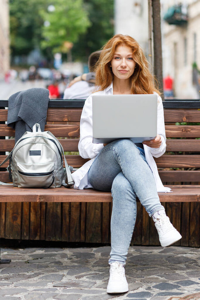 Jeune femme attrayante assise sur le banc à l'aide d'un ordinateur portable et regardant dans la caméra - Photo, image