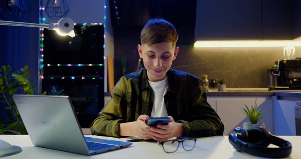 Successful teenage boy sitting at table using smartphone at home. Learning success, global education and e-leaning concept. - Fotografie, Obrázek