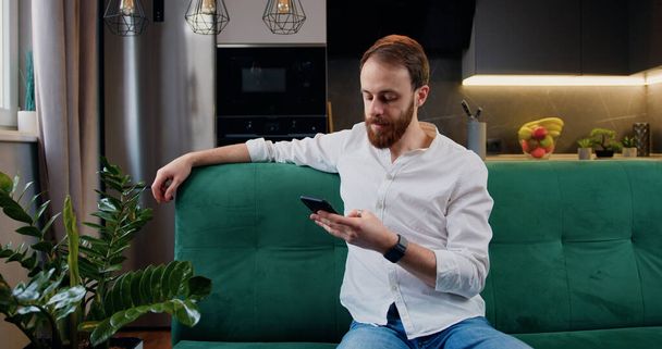 Portrait of man sitting on the sofa using phone in the modern kitchen texting message scrolling tapping technology. Caucasian male relaxing sitting on couch at home - Photo, Image