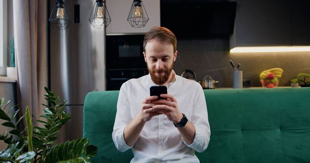 Handsome man browsing smartphone while sitting on sofa in kitchen at home. Cheerful bearded caucasian male in white shirt is texing and read a message. - Fotoğraf, Görsel