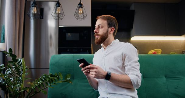 Attractive man typing message on smartphone sitting on sofa home. Electronic app use hold connection leisure lifestyle communication touching. - Foto, Imagen